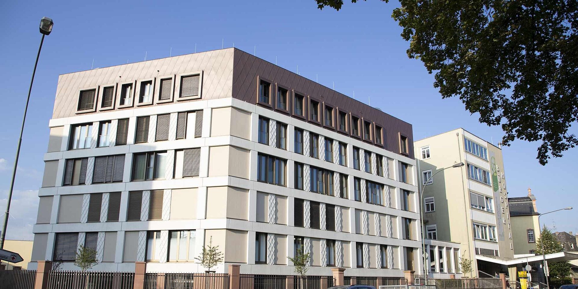 Ein modernes Krankenhausgebäude mit mehreren Stockwerken und großen Fenstern. Die Fassade kombiniert helle und dunkle Farbtöne, mit einem markanten Dachbereich. Im Vordergrund ist ein Baum mit grünen Blättern zu sehen, während der blaue Himmel im Hintergrund das Gebäude umrahmt.