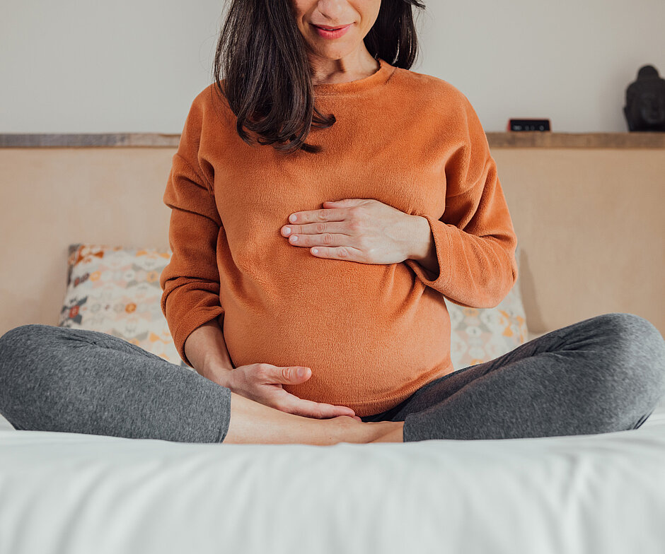 Schwangere Frau sitzt auf einem Bett und hält ihren Bauch (Schwangerschaft Guide)