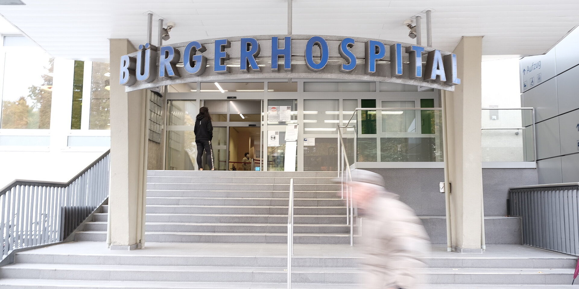 Der Haupteingang des Bürgerhospitals mit einem großen, gebogenen Schild über den automatischen Glastüren. Eine Person in dunkler Kleidung steht vor dem Eingang, während im Hintergrund weitere Personen im Inneren des Gebäudes sichtbar sind.