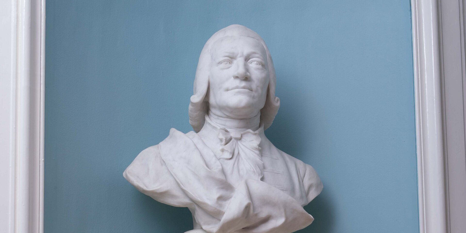 Eine weiße Marmorbüste eines Mannes mit schulterlangem Haar und Halstuch steht vor einer blau gestrichenen Wand. Die Statue zeigt ein ernstes Gesicht und ist auf einem Sockel montiert.
