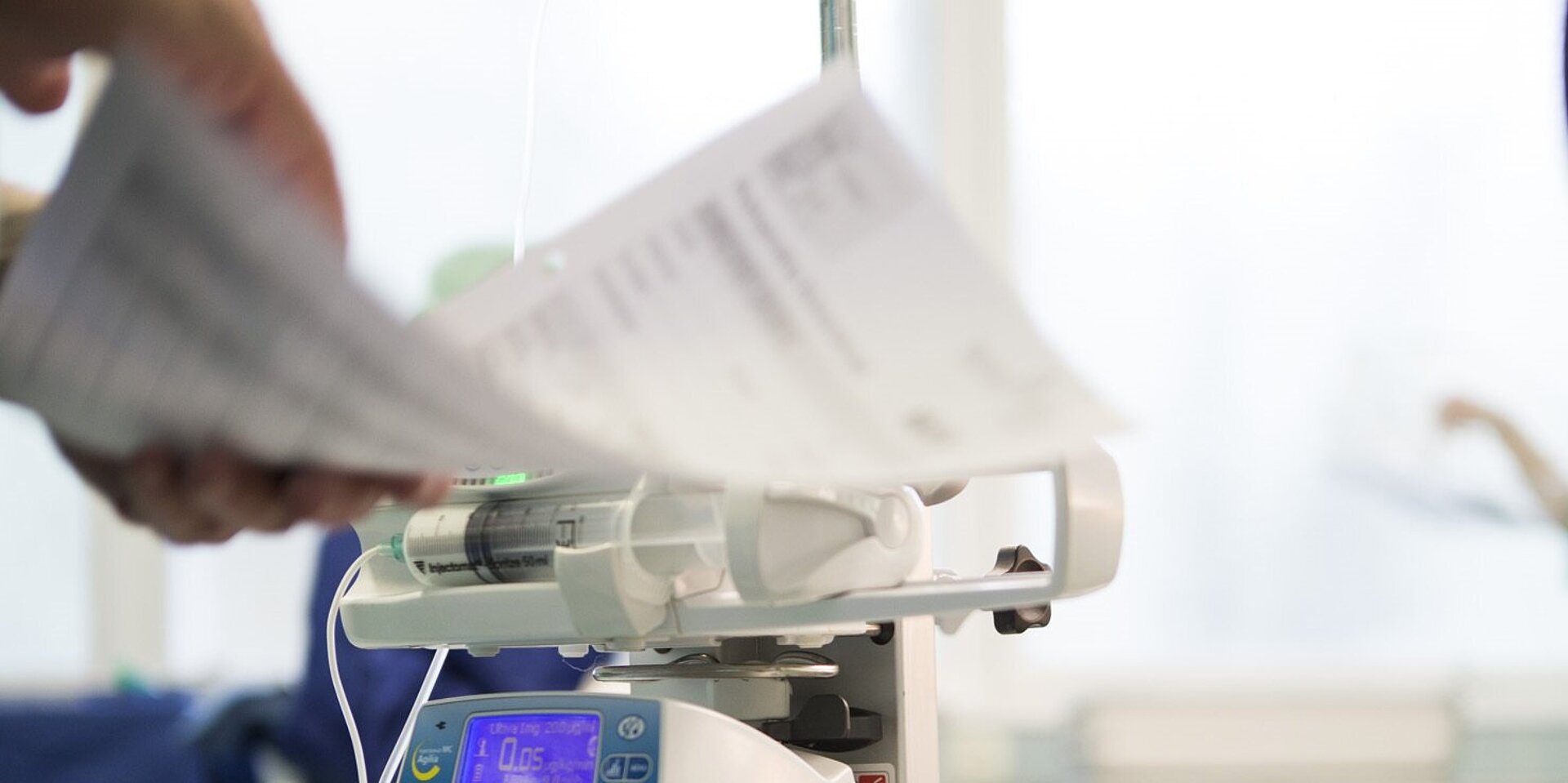 Ein medizinisches Infusionsgerät mit einer Spritzenpumpe wird bedient, während eine Person Patientendokumente überprüft.