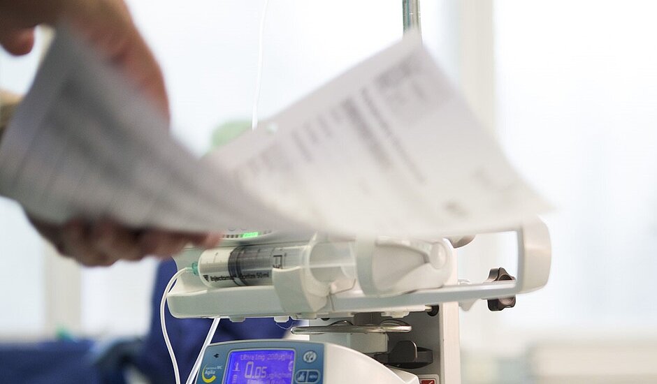 Ein medizinisches Infusionsgerät mit einer Spritzenpumpe wird bedient, während eine Person Patientendokumente überprüft.