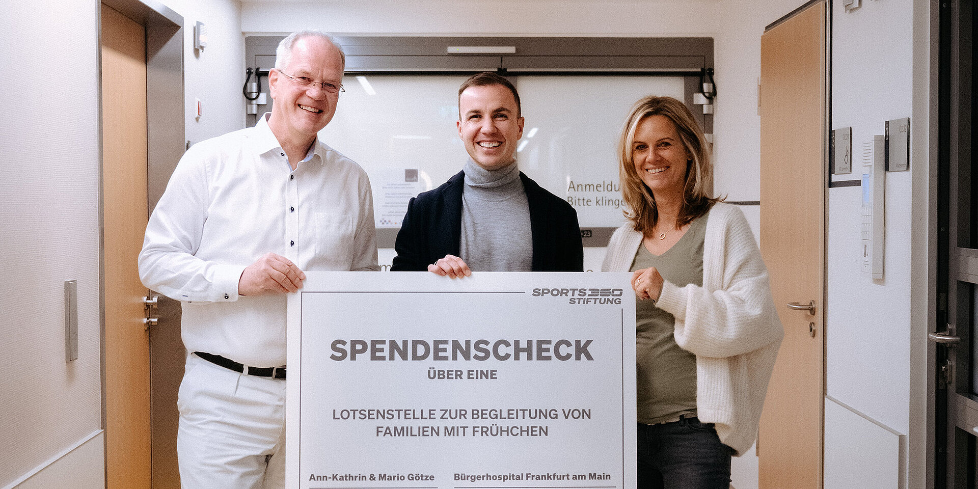 Drei Personen stehen in einem Krankenhausflur und halten gemeinsam einen großen Spendenscheck. Die Aufschrift des Schecks zeigt eine Spende für die „Lotsenstelle zur Begleitung von Familien mit Frühchen“. Alle drei lächeln und blicken in die Kamera.