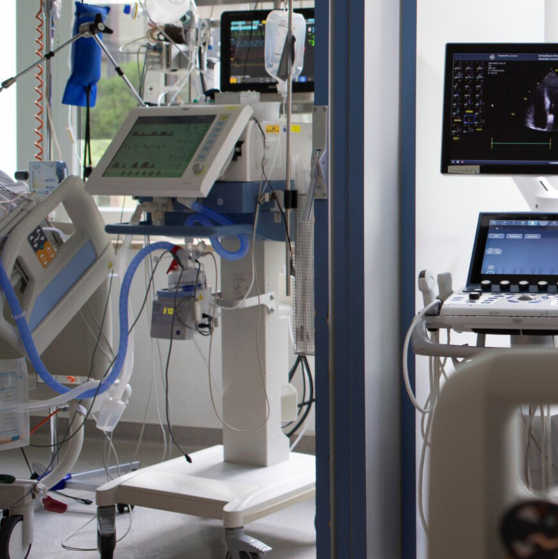 Ein Arzt in blauer OP-Kleidung untersucht einen Patienten im Krankenhausbett mit einem Stethoskop